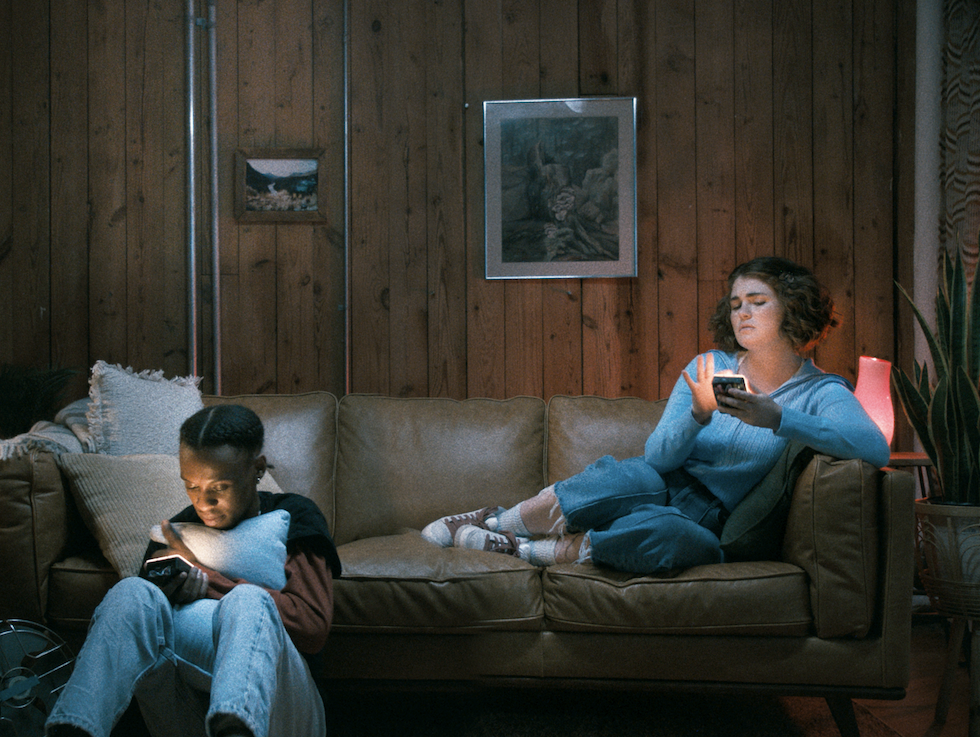 A girl and a guy sitting together, both holding their phones, looking at the screens with neutral expressions.