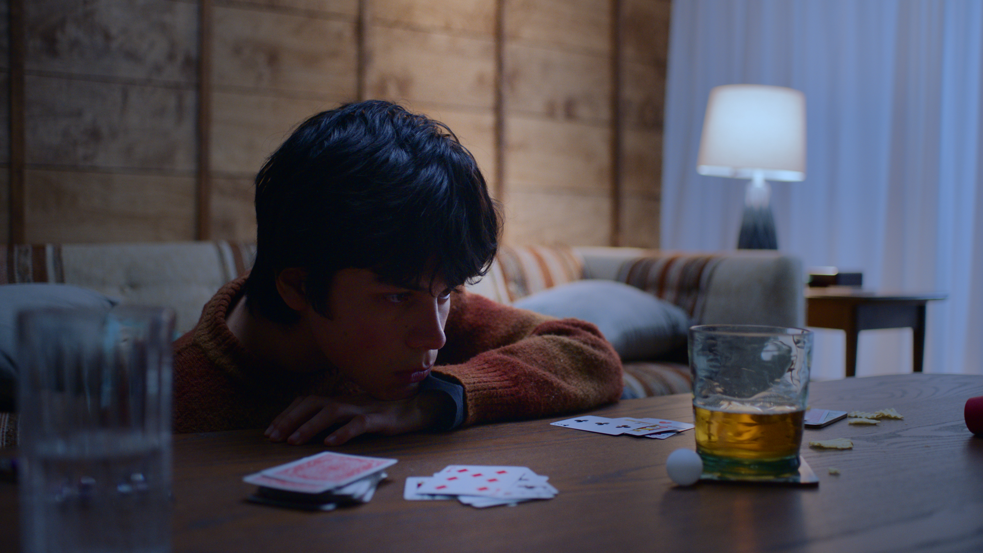 A guy staring at a glass with orange colored liquid on a table. The glass is next to a pile of poker cards.