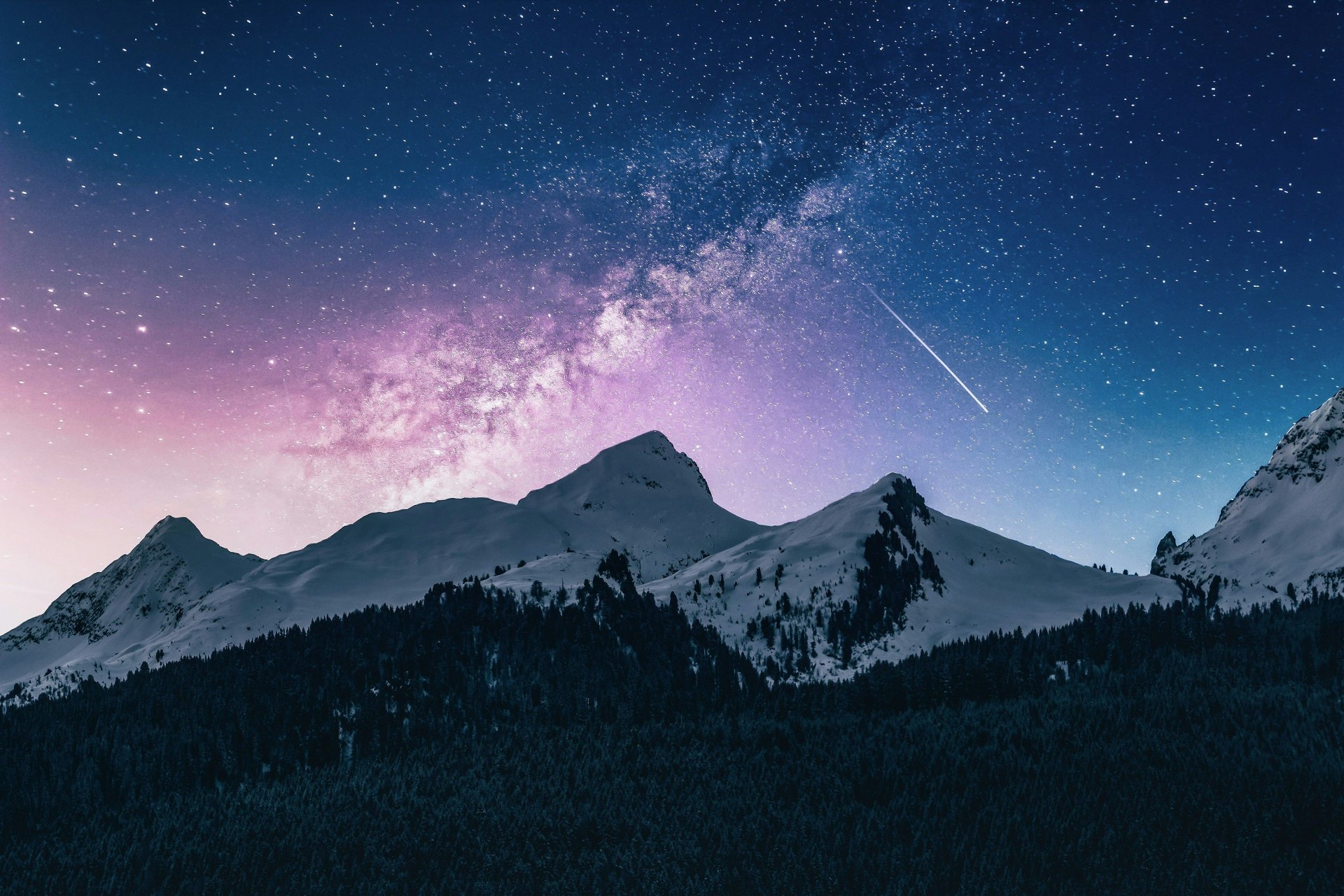 A star-filled night sky above snow-covered peaks and a dark alpine forest