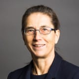 Elspeth Talbot Rice KC, smiling, in black suit and top, in front of a grey backdrop