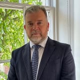 A headshot of His Honour Judge Unsworth KC wearing a suit and tie and standing in front of a window with trees outside