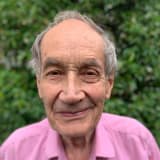 David Ainger smiling, in a pink shirt, standing in front of green foliage