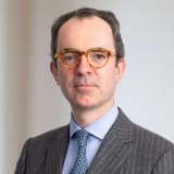 Headshot of a smiling David Scorey KC. He is wearing a grey pin striped suit jacket, blue tie and light blue shirt, and has on round tortoise shell glasses.