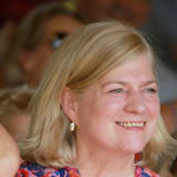 Her Honour Judge Whitehouse KC smiles in a crowd of people. Her blonde hair is shoulder length and she wears a blue and red floral shirt and small drop earrings