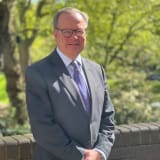 Malcolm Cree in a grey suit and purple tie, wearing glasses and smiling.