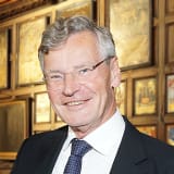 A smiling Michael Todd poses for the camera in front of coats of arms of dark wooden panels in the Great Hall of Lincoln's Inn. He wears a white shirt and dark suit and tie and has metal wire rimmed glasses
