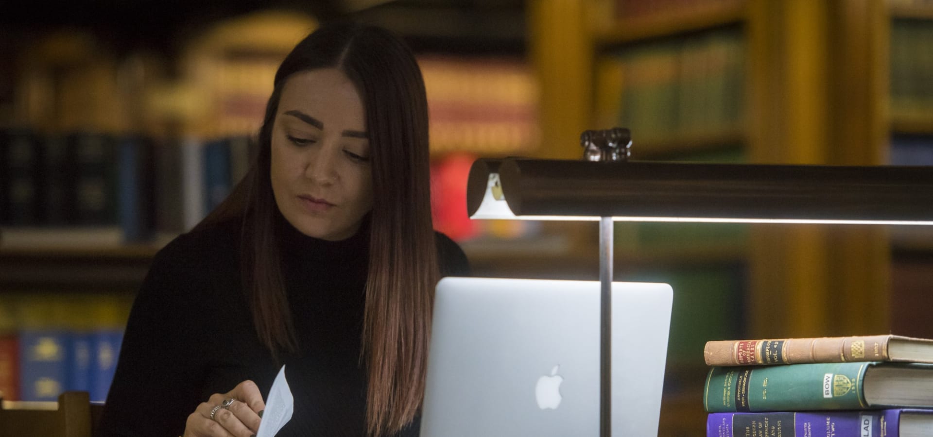 A women studying with books and a laptop in the Inn Library, a desk lamp in front of her