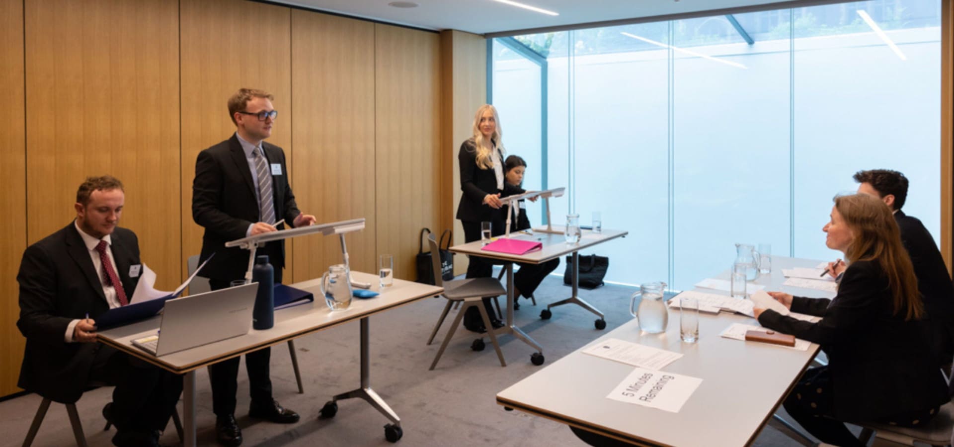 Five people in business attire participate in a mock trial. Two stand at lecterns presenting, while the remaining three sit at tables, listening intently.