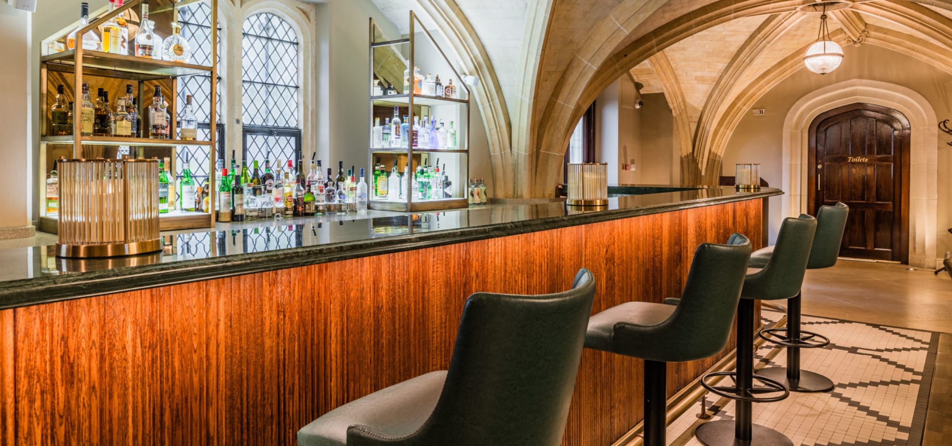 A stylish bar with a glossy countertop, illuminated wooden panels, and leather barstools. Arched windows and shelves of bottles create a warm, sophisticated ambiance.