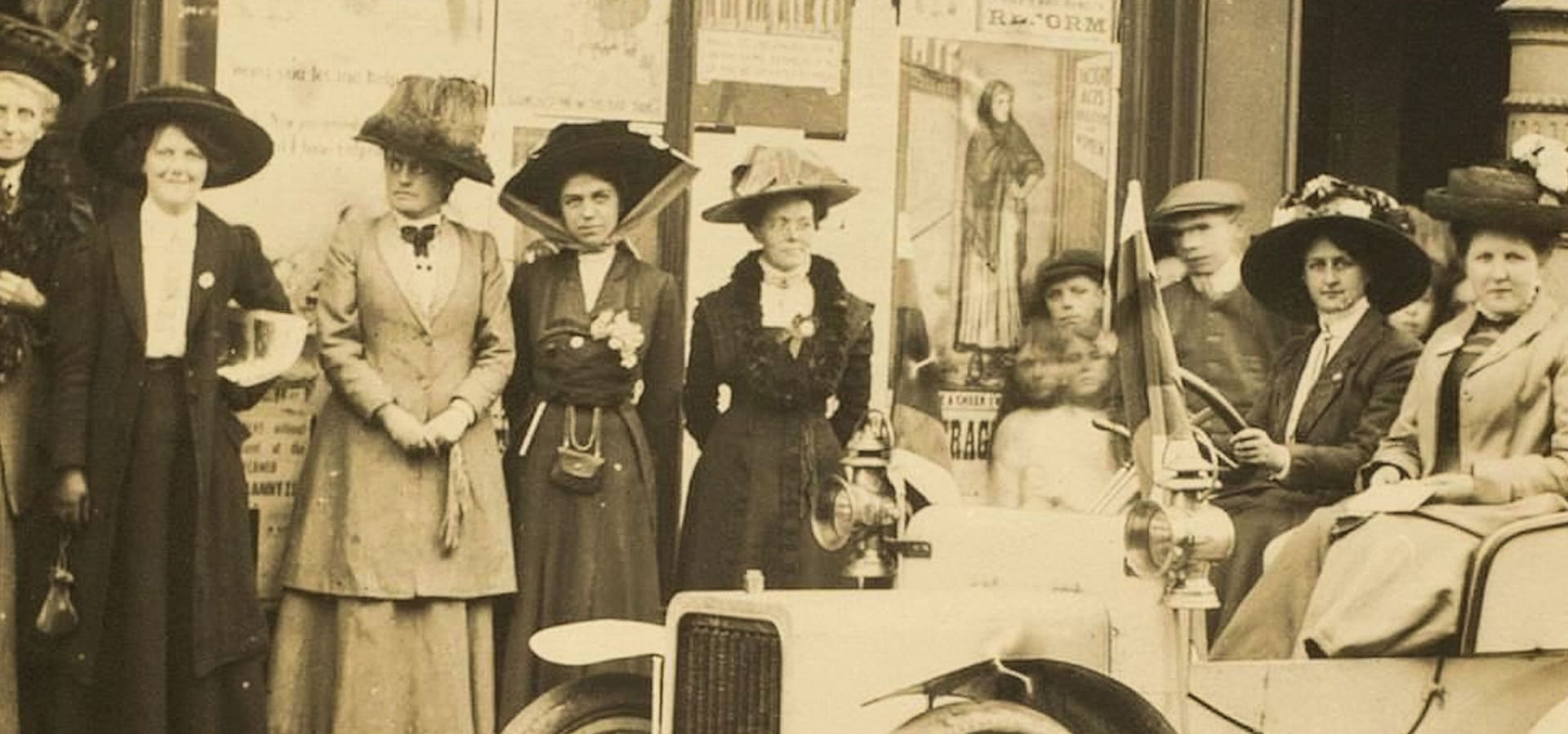 A sepia-toned photo of early 20th-century women suffragists, dressed in elegant hats and long dresses, gathered around a vintage car, exuding determination. Posters in the background hint at activism.