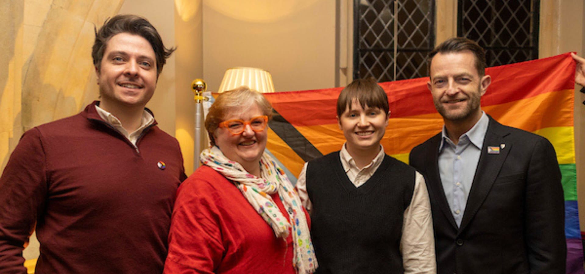 Four people stand smiling in front of a rainbow flag, signaling LGBTQ+ pride. They appear happy in a warmly lit room, creating an atmosphere of inclusivity.
