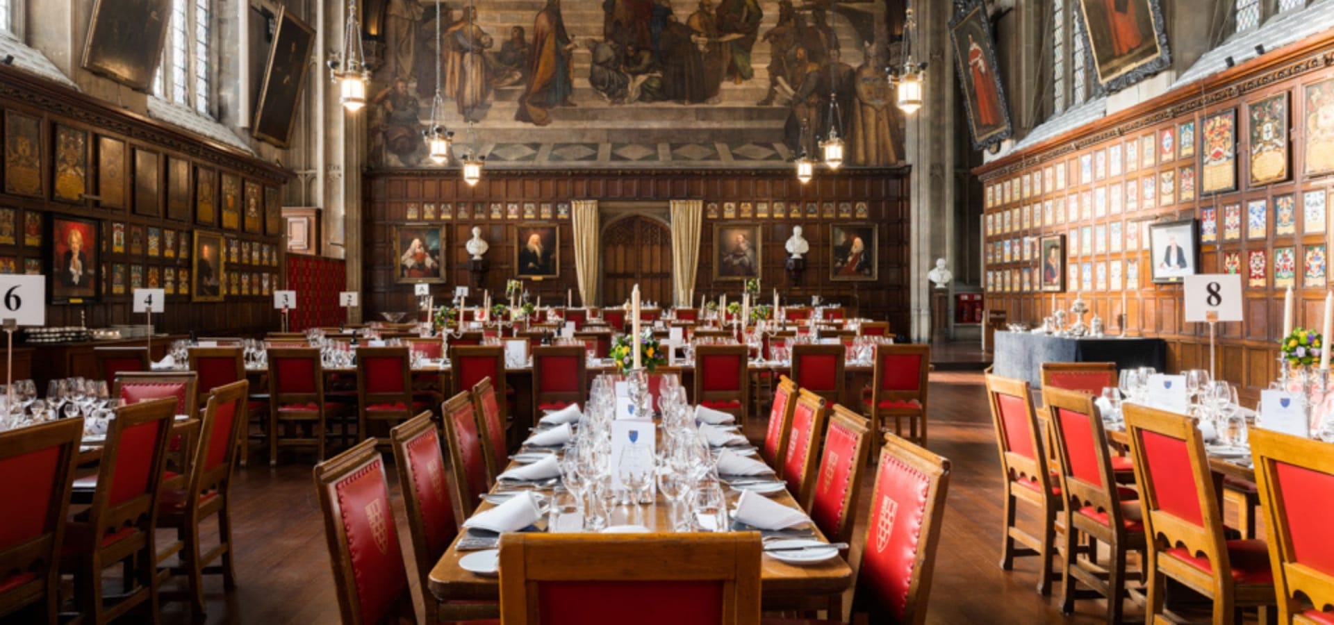 Grand dining hall with wooden tables and red high-backed chairs set for an event. Walls are adorned with portraits and ornate wood paneling, creating an elegant atmosphere.