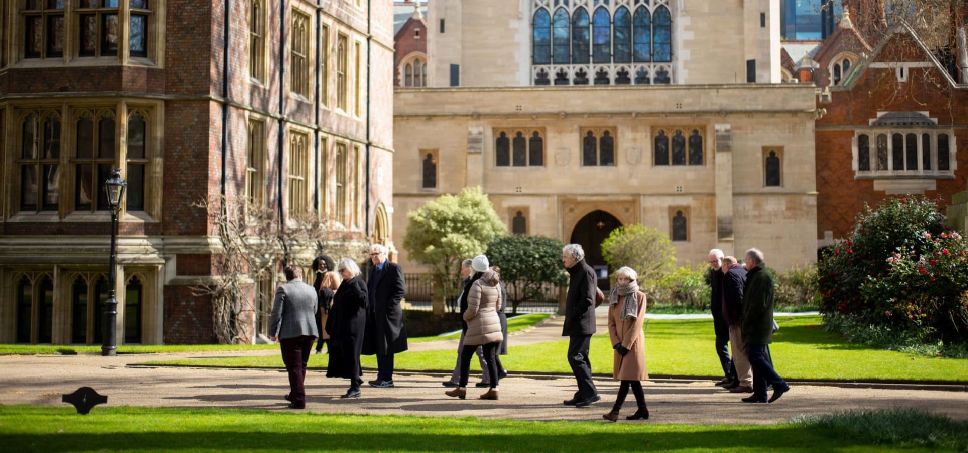 A group of people strolls through a sunlit, historic university campus with elegant, ivy-covered brick buildings and manicured lawns, evoking a sense of tradition.