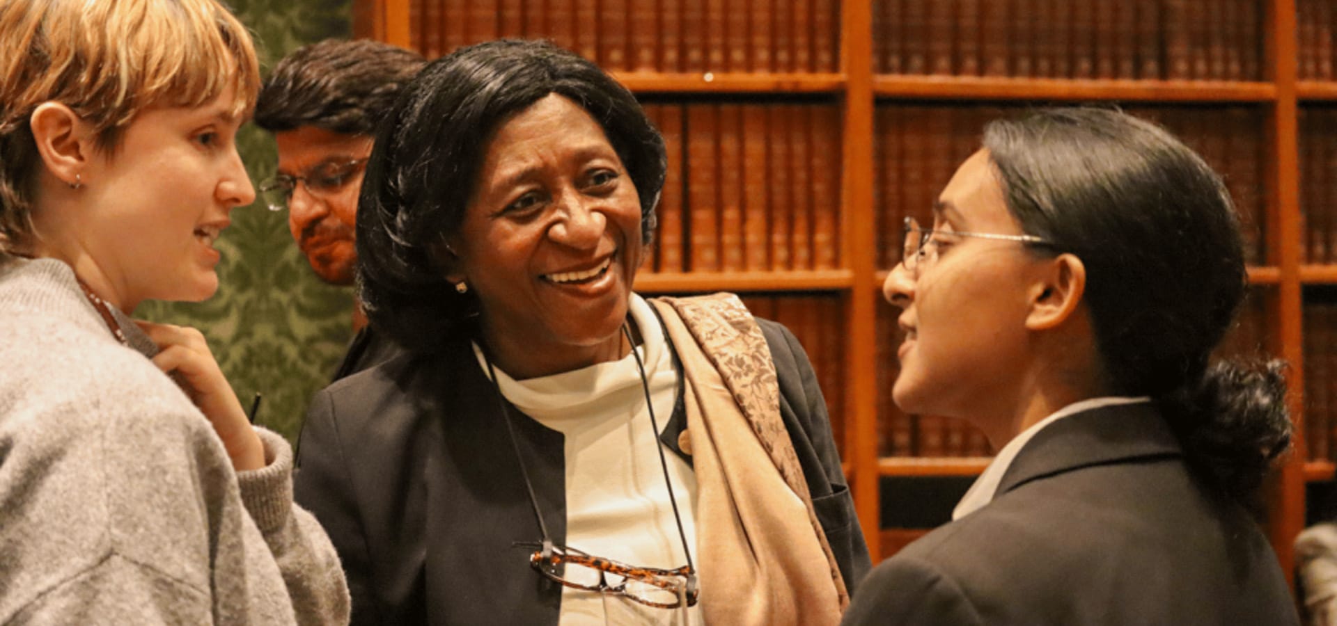 Three women converse and smile warmly in a library, with bookshelves behind them, creating a friendly, collegial atmosphere.