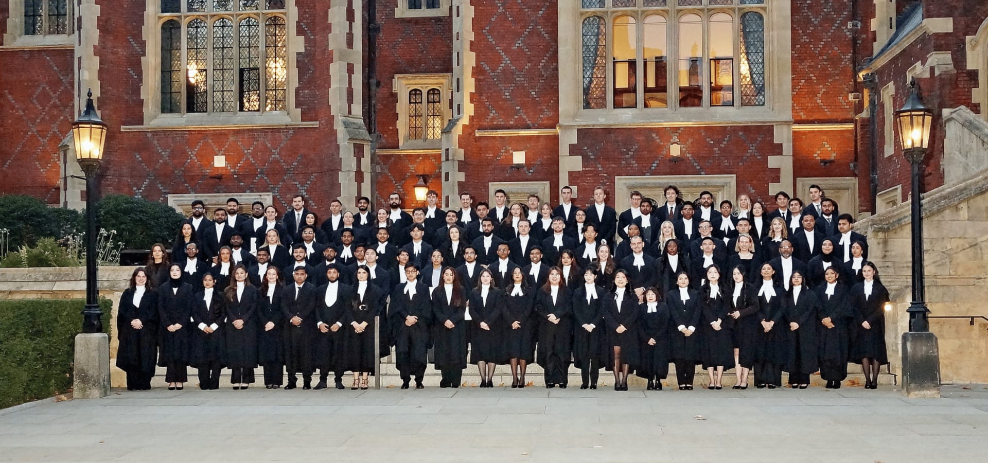 A group of around 100 newly called barristers, in professional attire, long black gowns, and white collars, pose for a group photo on the steps of Lincoln's Inn, in front of a large red brick Victorian building