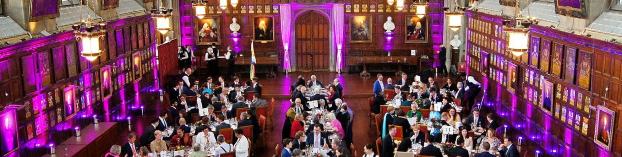 Full tables of people enjoying the Pride Diversity Dinner at Lincoln's Inn. People are dressed in colourful smart attire and the hall is lit with purple lighting. Their are Pride flags on tables.