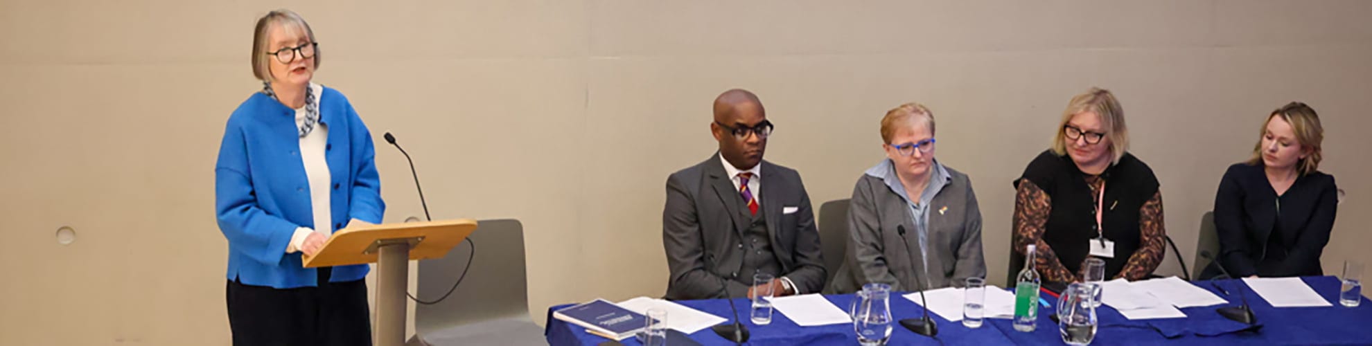 A woman in a blue cardigan speaks at a podium. Beside her, four people sit at a table with papers and water glasses, listening attentively.
