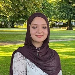 Bibi Khan headshot - she wears a brown head covering and is standing on a grassy lawn in the sunshine