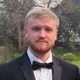 James Woods, in black-tie, standing in front of tree and flowers. He has blond hair and strawberry blond beard and mustache