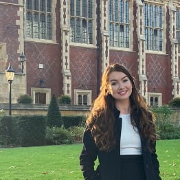 Jennifer White stands and smiles on the Benchers' Lawn in front of Lincoln's Inn,. She has ling brown hair and is in a white top and black jacket and skirt.