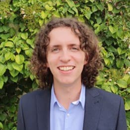 Joe Norris haedshot. He is smiling, has shoulder length curly brown hair and wears a blue suit. He is in front of green leafy foliage.
