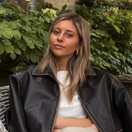 Mille Thompson sittign in a rattan chair in front of greenery. She has long blond hair and wears a white jumper with a leather jacket resting on her shoulders.