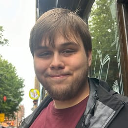 Nathan Lynas smiles into the camera. He is outside a bookshop and have a light brown, short beard and wears a red t-shirt.