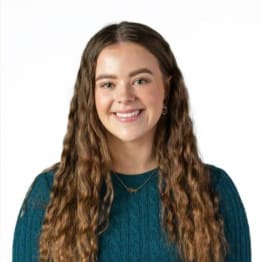 Rhiannon James smiles broadly. She as very long wavy brown hair and is in a teal jumper. She is in front of a plain white background.