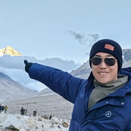 Steven, wearing a thick winter jacket, woolly hat, and aviator sunglasses, stands in a mountain range, pointing at a snowy peak in the distance.