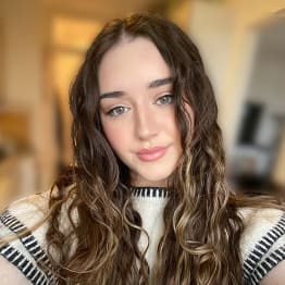 Ursula O'Kane selfie. She has long brown wavy hair and wears and white jumper with black stitching. The background is in soft focus.