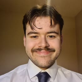 William Lewis, in a white shirt and navy blue tie, smilesto the camera. He has a brown mustache and stubble.