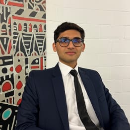 Zarrar Haider Bhatti sit in front of an abstract art work on a white wall. he is in a black suit and tie and wear black framed glasses.