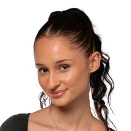 Smiling young woman with dark, curly hair in a high ponytail, small earrings, and a dark top against a plain background, conveying a friendly look.