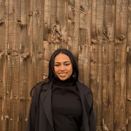 A woman in a black outfit stands smiling against a rustic wooden wall. Her relaxed expression and the warm tones create a friendly, inviting mood.