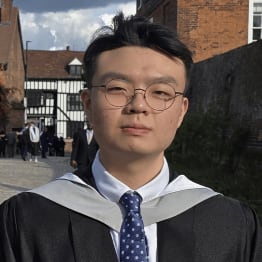 A young graduate with glasses and a blue polka dot tie stands confidently in a black gown. Background includes brick and timber buildings on a sunny day.
