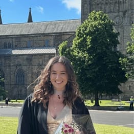A smiling graduate in a black gown holds a bouquet in front of a historic stone building and lush trees under a clear blue sky, conveying joy and achievement.