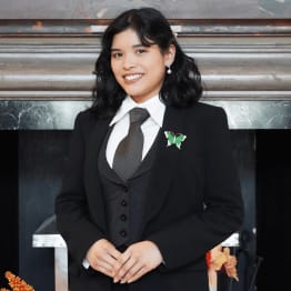Smiling person in a black suit with a white shirt and tie, wearing a butterfly brooch, stands confidently in front of a dark, textured background.