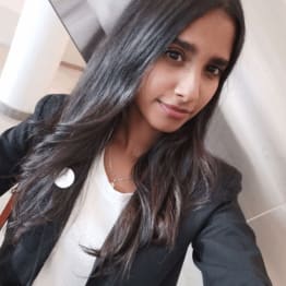 A woman with long dark hair smiles slightly, wearing a black blazer and white top. She sits indoors against a light-colored wall, conveying a professional tone.