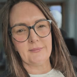 A woman with long brown hair and large glasses smiles gently at the camera. The background is softly blurred, conveying a calm and approachable tone.