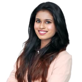 Smiling woman with long dark hair wearing a light pink blazer poses confidently with arms crossed against a white background, conveying professionalism.