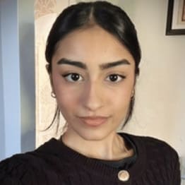 Woman with dark hair pulled back, wearing a black patterned sweater, looks into the camera against a light indoor background. She has a calm expression.