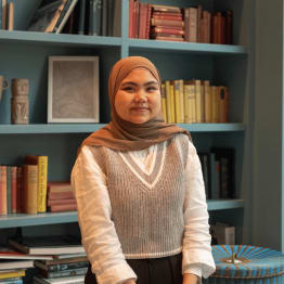 Young woman in a brown hijab and grey sweater vest stands smiling in front of a bookshelf filled with colorful books, conveying a warm and inviting tone.