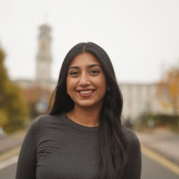 Smiling woman with long dark hair stands on a blurry street with trees and a tower in the background. She wears a gray top, conveying warmth and confidence.