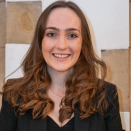 Smiling person with long brown hair, wearing a black blazer. Background features a textured, wooden wall. The tone is warm and professional.