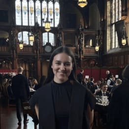 A smiling woman in a formal black outfit stands in an ornate hall with stained glass windows and chandeliers. People sit at tables having a formal meal.