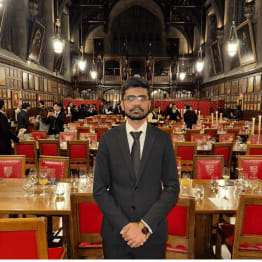 A person in a black suit stands in an elegant, historic hall with high ceilings, chandeliers, and red-cushioned chairs. The atmosphere is formal and grand.