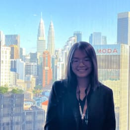 A woman in glasses and a blazer smiles in front of a window. The city skyline behind her features tall buildings, including two prominent towers.