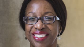 Headshot of a smiling Nicola Williams. She is a Black woman, who wears her straightened hair swept back behind her in a middle parting. On her face there are dark, rectangular glasses. She wears an orange top and a patterned cardigan that features speckles of burgundy, red and white.
