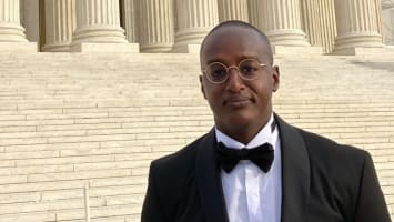 Samuel Buyoya, a clean-shaven Black man with short hair and round gold wire-rimmed glasses, dressed in Black Tie, stands on stone steps in front of large Romanesque pillars