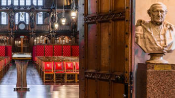 The Great Hall Historic with ornate woodwork and red upholstered chairs, flanked by large wooden doors. There are two marble busts on pedestals.
